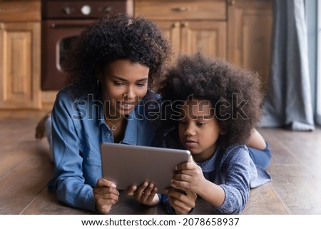 Happy young African ethnicity mother using digital computer tablet with little biracial kid daughter, watching cartoons funny photo video content online, lying on warm floor, tech addiction.