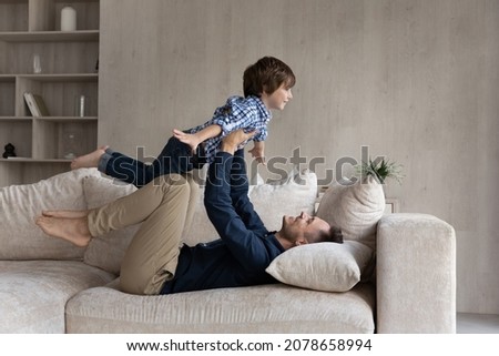 Dad lying on couch lifts on outstretched hands little 6s son. Happy cute boy play plane fly in air while his father raise him up on arms. Active weekend at home, dreams about holiday, leisure concept Royalty-Free Stock Photo #2078658994