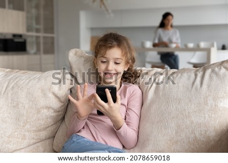 Smiling little girl having fun with smartphone while mother cooking on background, cute kid looking at phone screen, playing mobile device game, watching cartoons, sitting on cozy couch at home
