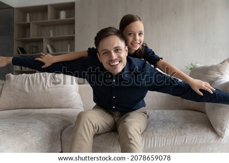 7s daughter piggyback dad, sit on sofa having fun raise their arms like plane wings play together at home look at camera, spend time enjoy playtime in living room. Happy Father Day, fatherhood concept