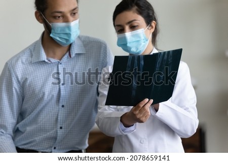 Indian woman doctor in protective medical mask holding x-ray image, consulting African American man patient at meeting in hospital, young female physician surgeon examining bones, checkup concept