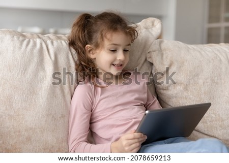 Close up curious adorable cute girl kid using tablet, looking at electronic device screen, sitting on cozy couch at home alone, smiling little child playing game, having fun online, watching cartoons