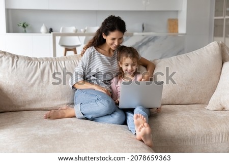 Smiling mother with adorable daughter using laptop sitting on couch together, happy family young mom and cute girl kid looking at computer screen, chatting online by video call, watching cartoons