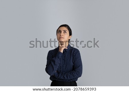 Thoughtful young 30s Indian ethnicity businesswoman touching chin, looking up in distance, considering problem solution, feeling doubtful isolated alone on white wall or grey studio background. Royalty-Free Stock Photo #2078659393