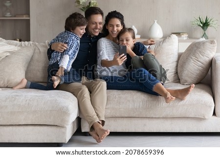 Family enjoy funny mobile app relaxing on couch. Latin couple and preschool children use smartphone watch videos on internet laughing have fun spend weekend at home with online amusing virtual content Royalty-Free Stock Photo #2078659396