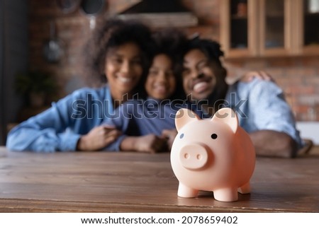 Close up piggybank standing on table with blurred happy African American couple parents and small child daughter on background. Friendly biracial family planning future investment or saving money. Royalty-Free Stock Photo #2078659402