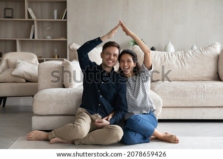 Happy Hispanic 35s couple joining hands under heads showing roof symbol sit on warm floor in fashionable living room. Homeowners spouses at relocation day, bank loan for young family, tenant concept Royalty-Free Stock Photo #2078659426
