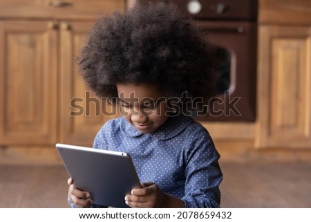 Happy cute small 6s African ethnicity child girl playing online games on touchpad gadget, watching entertaining photo video content in social networks or using educational apps, tech addiction.
