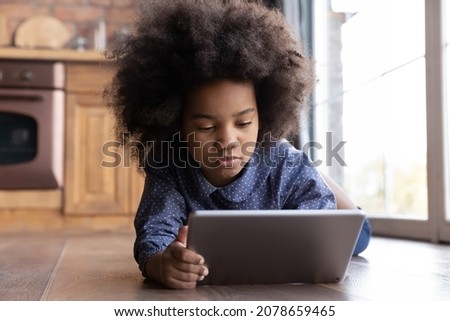 Concentrated adorable little African American kid girl playing online games on digital computer tablet gadget, lying on warm heated floor, watching entertaining photo video content in social networks.