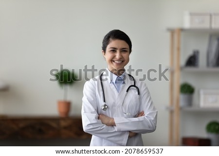 Head shot portrait smiling Indian woman doctor with folded hands standing, confident young female physician general practitioner therapist wearing white uniform with stethoscope looking at camera