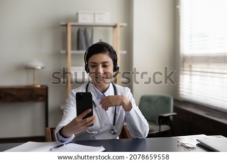 Smiling Indian woman doctor in wireless headphones looking at smartphone screen, talking, young female physician general practitioner explaining, consulting patient by video call in office