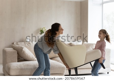 Happy mother with little daughter carrying armchair, smiling young mom and cute girl child decorating cozy living room in new own home apartment, moving day and relocation, renovation concept Royalty-Free Stock Photo #2078659567