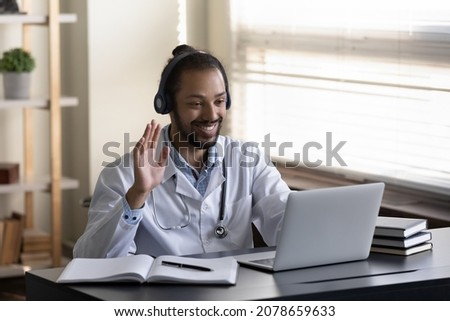 Smiling young African American man doctor in wireless headphones using laptop, greeting client, waving hand at computer webcam, consulting patient by video call online, engaged in virtual meeting