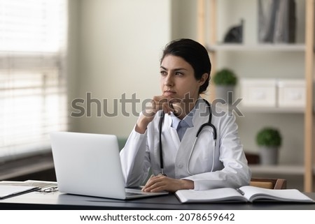 Thoughtful serious Indian female doctor physician touching chin sitting at work desk in office with laptop, looking in distance, pensive practitioner in white uniform with stethoscope thinking Royalty-Free Stock Photo #2078659642