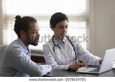 Indian woman doctor in uniform with stethoscope consulting African American patient, using laptop, showing discussing medical checkup results or symptoms, looking at computer screen in office