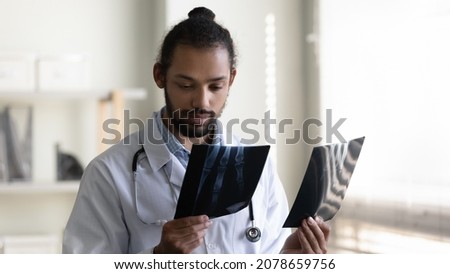 Serious young African American man doctor surgeon in white uniform with stethoscope holding x-ray images, analyzing patient medical checkup results, standing in hospital office, healthcare concept