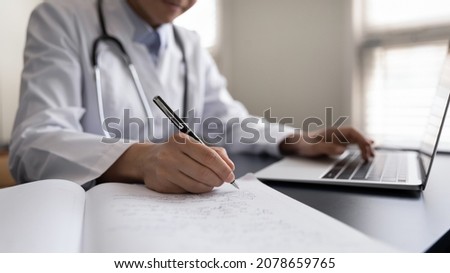 Crop close up Indian woman doctor in white uniform with stethoscope taking notes, using laptop, writing in medical journal, professional therapist practitioner filling documents or patient card Royalty-Free Stock Photo #2078659765