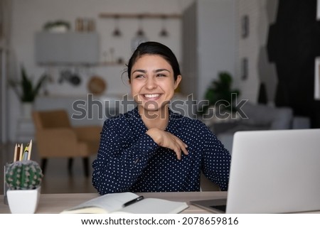 Smiling dreamy young Indian ethnicity woman sitting at table in modern home office, thinking of new creative online project or received email with pleasant news, considering problem solution.