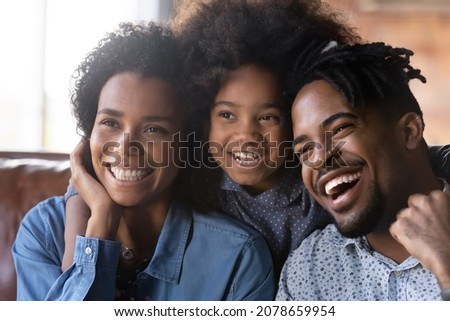 Head shot happy bonding African American two generations family parents resting with little cute child daughter on cozy sofa, looking in distance, watching funny smart TV programs or shows at home.