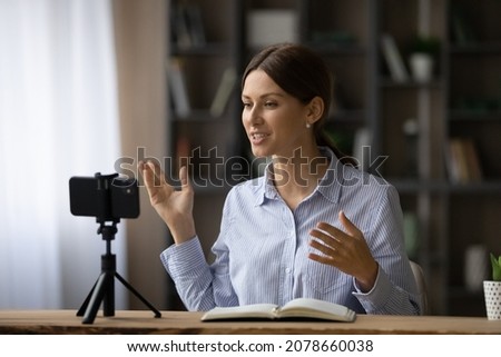 Attractive professional young 30s woman speaker sit at desk talk to internet audience through streaming video conferencing app, record educational webinar on cellphone. Coaching, modern tech concept Royalty-Free Stock Photo #2078660038