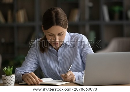 Concentrated serious young woman sit at desk near laptop jotting information in notebook. Making important notes, write personal records to daybook, keep a diary, create business to-do list concept Royalty-Free Stock Photo #2078660098