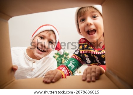 Happy surprised Caucasian little boys in Santa's hat looking inside a cardboard box, at home near Christmas tree, bottom view. Excited little children opening Christmas present. 