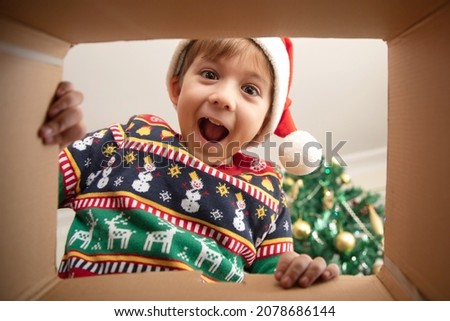 Happy surprised Caucasian little boy in Santa's hat looking inside a cardboard box, at home near Christmas tree, bottom view. Excited little kid opening Christmas present. 