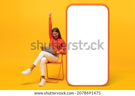 Winner. Excited young woman sitting on chair with her laptop and huge phone mock up with blank screen, shouting yes, isolated on yellow 