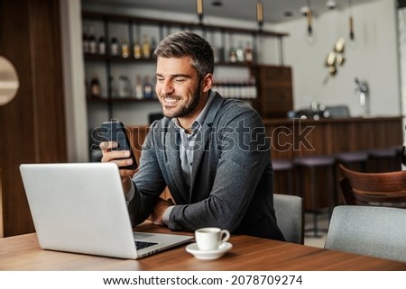 A happy businessman is sitting in a coffee shop and checking on his bank account on the mobile. There is a laptop on a table. A man using the phone for e-banking Royalty-Free Stock Photo #2078709274