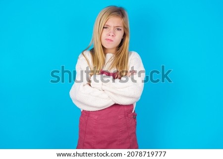 Picture of angry caucasian little kid girl wearing jumpsuit over blue background crossing arms. Looking at camera with disappointed expression.