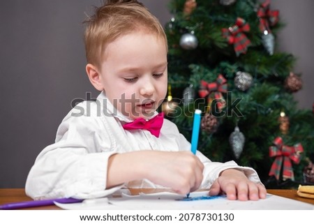 Lovely kid in white shirt with bow tie is writing letter with wishes to Santa or painting greeting card for parents to Christmas with colored pencil. House is decorated for holidays.
