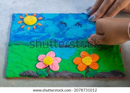 Children's clay sculptures depict a landscape with flowers in front and fields, sky, mountains and sun in the background.