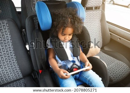 Little African-American girl with mobile phone watching cartoons in car safety seat