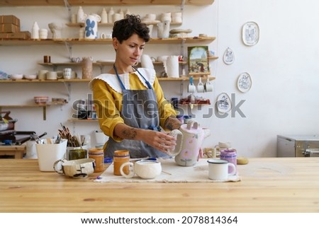 Artistic pottery master at work: happy woman in apron holding handmade jug working in ceramics studio. Craftswoman creating handicraft crockery in workshop. Craftsmanship and entrepreneurship concept Royalty-Free Stock Photo #2078814364