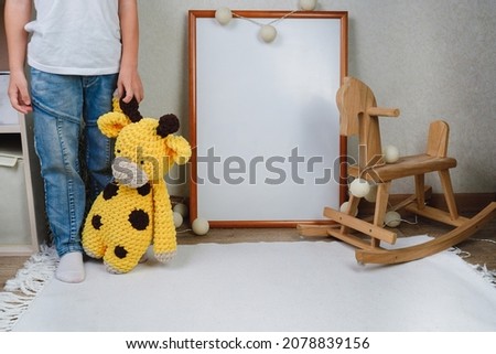 The child is holding a soft toy. Mockup of the picture in the children's room. wooden horse and knitted toys. The interior of the nursery in beige colors.