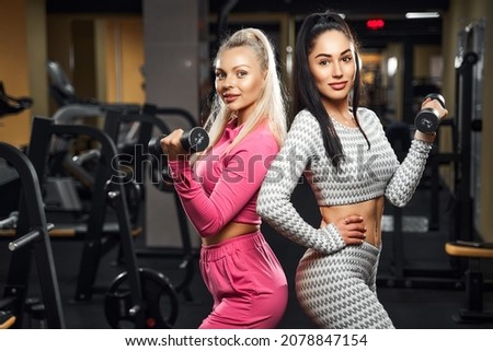 Two beautiful Sports girls doing exercises for biceps with dumbbells while standing and posing in gym. Fitness concept. Perfect body 