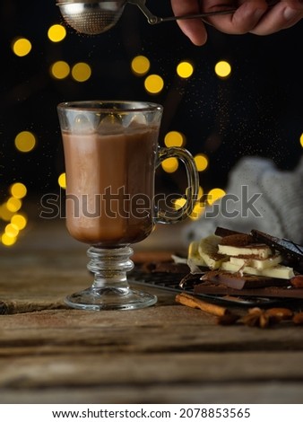 Festively served coffee table for one person. Large glass cup of coffee with milk, pieces of chocolate. Golden lights shine on a black background. Holiday, New Year, Christmas spirit.