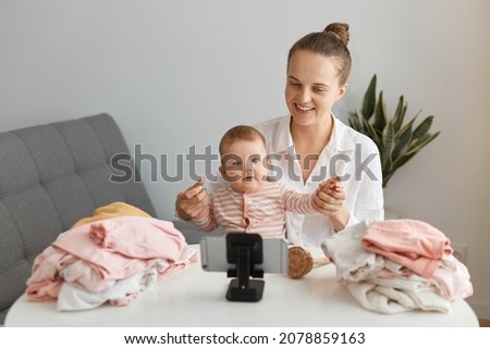 Smiling positive young adult woman blogger sitting in front of phone on tripod and shooting video, posing with her infant child at table and looking at device display.