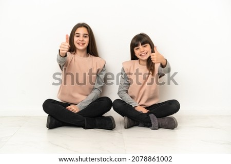 Little sisters sitting on the floor isolated on white background giving a thumbs up gesture because something good has happened