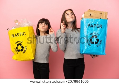 Little sisters recycling paper and plastic isolated on pink background with fingers crossing and wishing the best