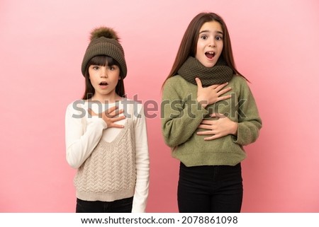 Little sisters wearing a winter clothes isolated on pink background surprised and shocked while looking right