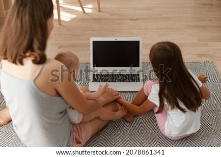 Back view of dark haired mother and daughter sitting on the living room floor and looking at laptop display, copy space for promotional text or your advertisement.