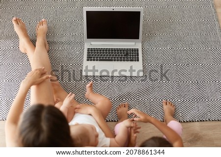 Top view portrait of mom and her daughters sitting on the living room floor and watching cartoons or funny videos via laptop, notebook with blank screen for your advertisement.