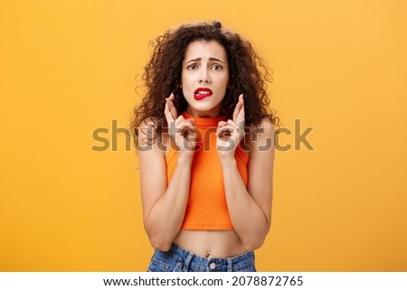 Worried and anxious silly caucasian female with curly hair in red lipstick and cropped top biting lower lip nervously looking concerned crossing fingers for good luck making wish over orange wall