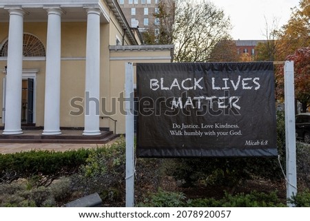 Black lives matter sign with scripture from bible in front of yellow church with white pillars in public street in Washington DC, USA. No visible people. Tall buildings in background.