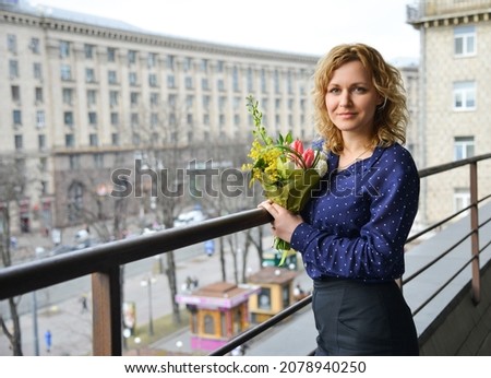 Smiling woman in a blue blouse with a bouquet of flowers stands on the balcony