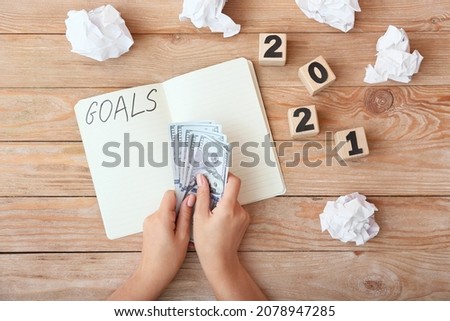 Female hands with money, notebook with text GOALS and cubes with 2021 on wooden background