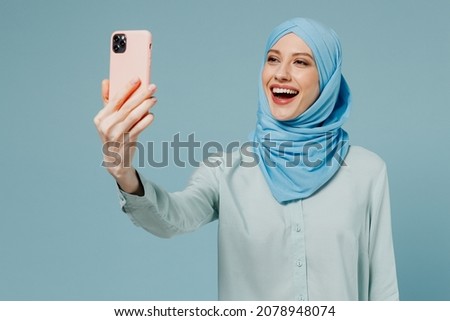 Young smiling happy arabian asian muslim woman in abaya hijab doing selfie shot on mobile cell phone isolated on plain blue background studio portrait People uae middle eastern islam religious concept