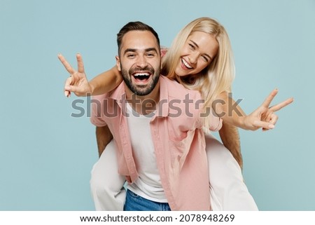 Young happy fun couple two friends family man woman in casual clothes giving piggyback ride to joyful, sit on back show v-sign gesture together isolated on pastel plain light blue background studio