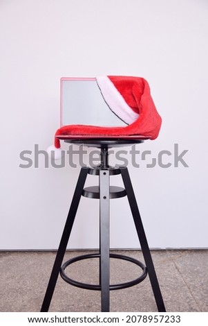 A red laptop decorated with a New Year's santa claus hat stands on a black metal chair on a white background